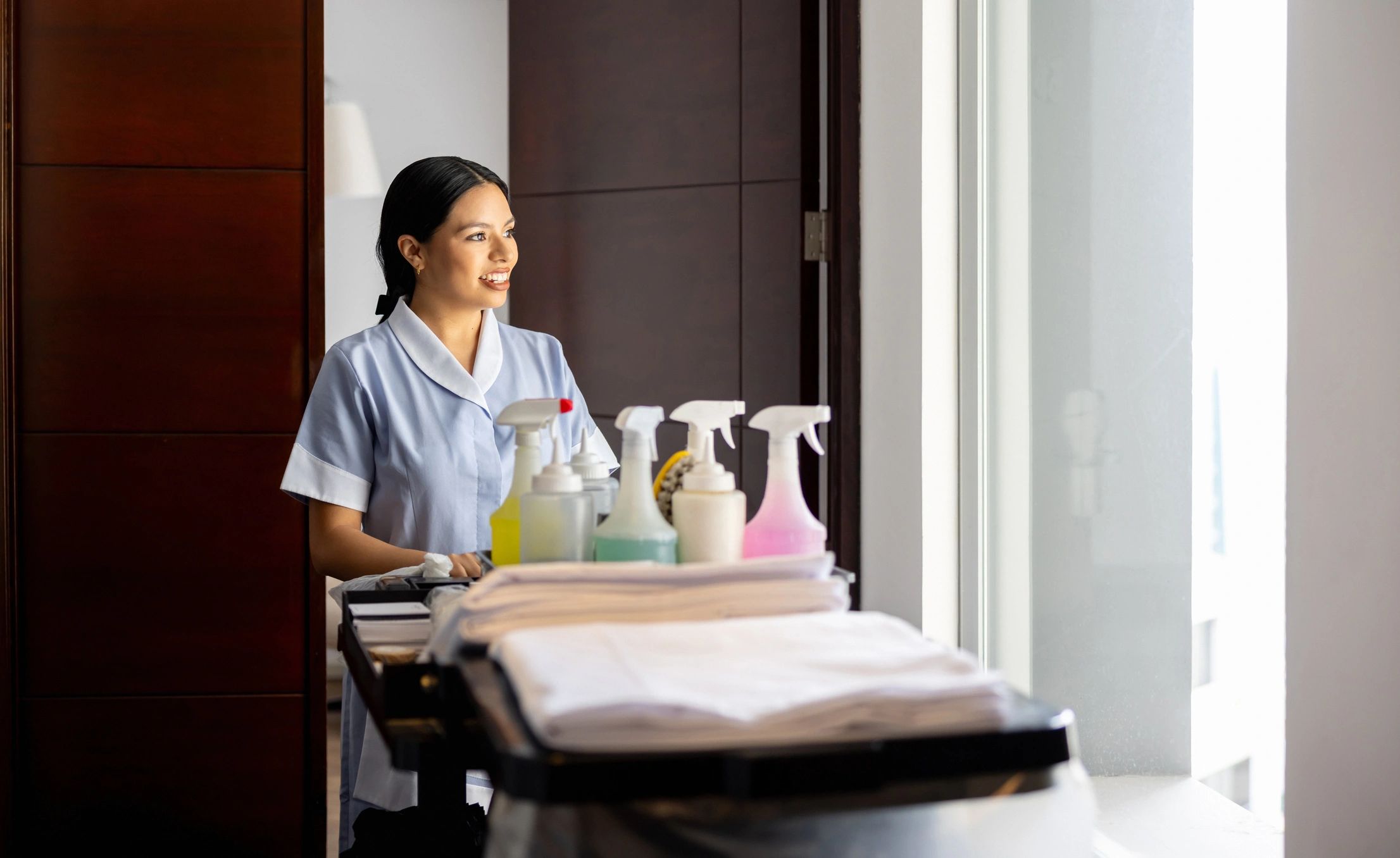 A hotel housekeeper prepares a room with cleaning supplies on a cart.