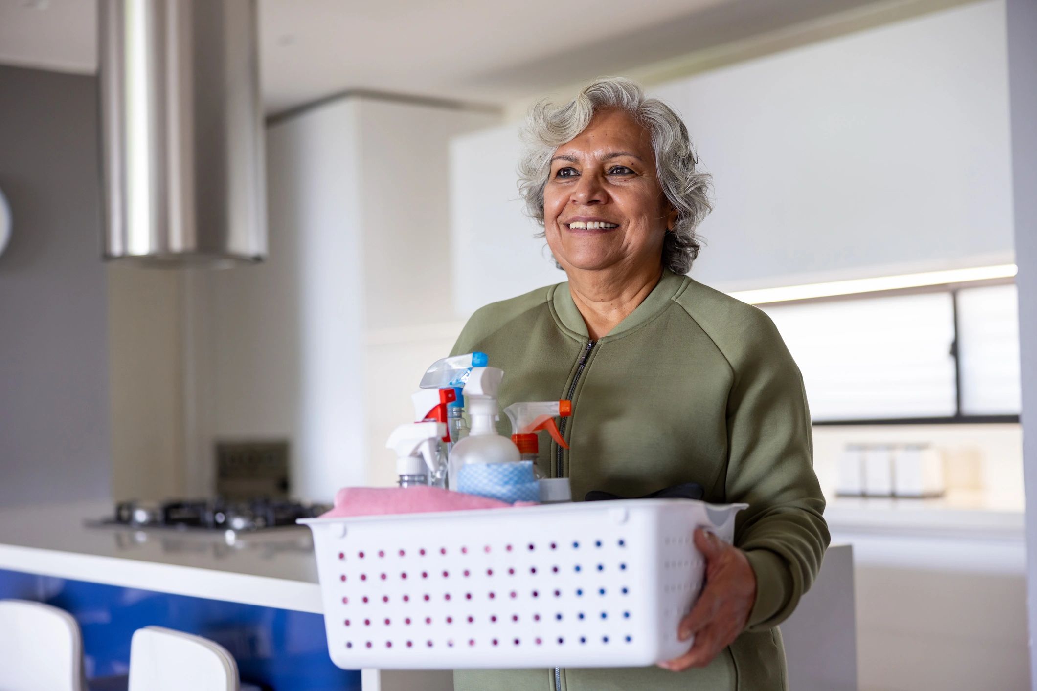 Smiling elderly woman holding a laundry basket with cleaning supplies.