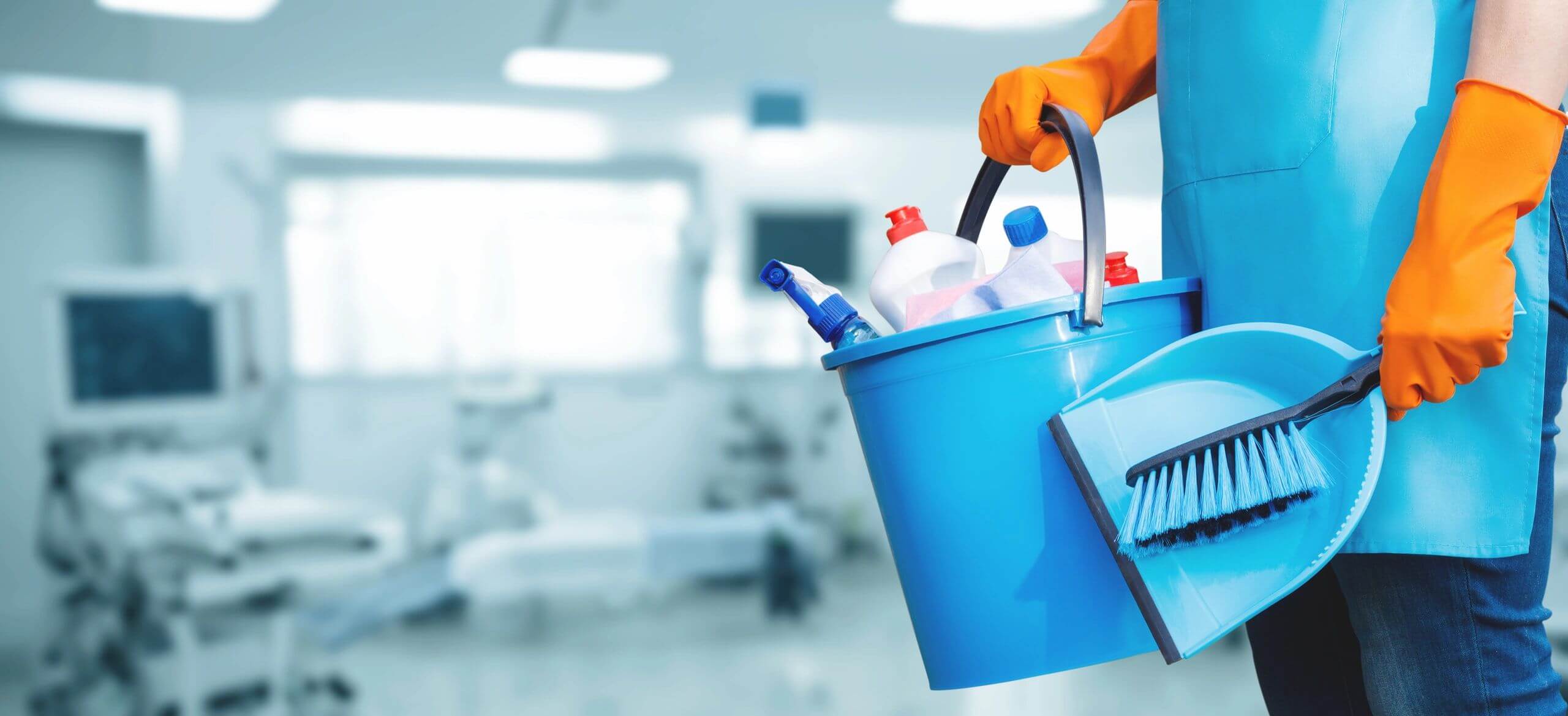Hand holding a blue bucket filled with cleaning supplies in a bright room.
