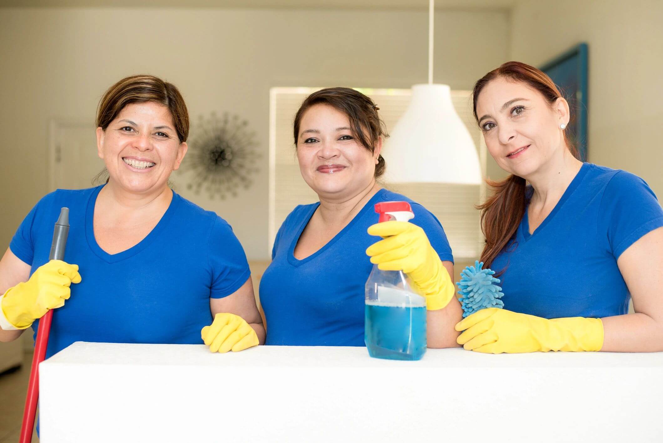 Three women in blue shirts cleaning with gloves and spray bottle.